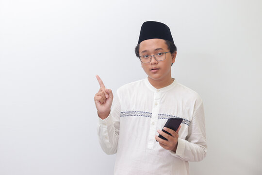 Portrait of young excited Asian muslim man pointing away with his finger while using mobile phone. Isolated image on white background