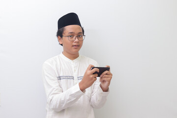Portrait of young Asian muslim man holding a smartphone and playing mobile game. Isolated image on white background
