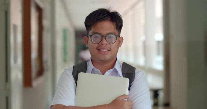  A happy smile of an Asian male teenage high school student wearing thick glasses. wearing a white sweater, carrying a laptop with a backpack standing on the corridor in the building.