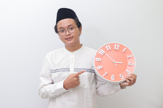 Portrait of young attractive Asian muslim man pointing and holding a clock. Isolated image on white background