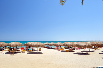 Straw shade umbrellas on sea tropical beach with resting sunbeds against blue vibrant sky in summer