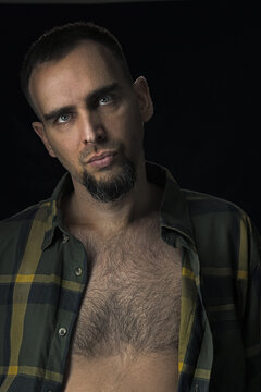 Handsome man in an unbuttoned checked shirt exposing his hairy torso. Portrait of a bearded Caucasian man against a black background. Sexy man looking at the camera.