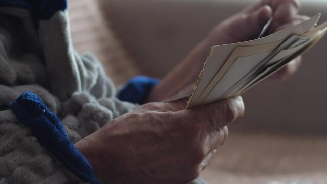 a pensioner sitting on a sofa in home clothes holds old photos in his hands. an elderly man examines the family archive. the concept of nostalgia, memories of the past, elderly people at home
