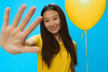 Asian woman holding a yellow balloon on a blue background in a yellow T-shirt smiling with teeth,...