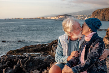 Lovely senior couple exchange a kiss sitting on the rocks at sea enjoying sunset light. Romantic relaxed lifestyle for a caucasian couple of retirees