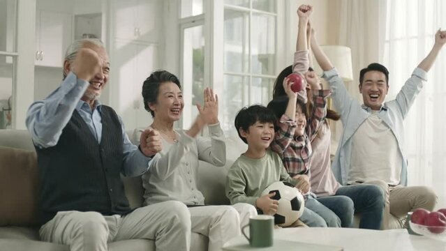 happy three generation asian family sitting on couch at home watching soccer match on tv and cheering