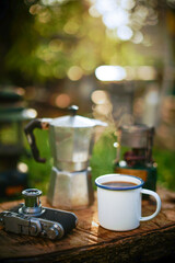 Black coffee in a white enamel mug on old wooden background with a film camera and coffee equipment Camping in the forest in the morningม selective focus, soft focus.