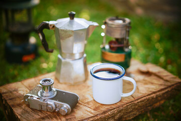 Black coffee in a white enamel mug on old wooden background with a film camera and coffee equipment Camping in the forest in the morningม selective focus, soft focus.