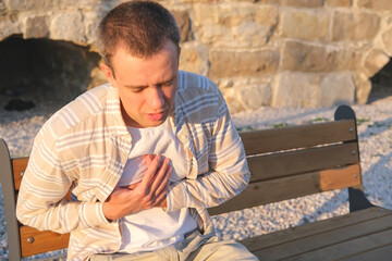 Handsome man has a heart pain on the street, person folds his palms on the chest and sits on the bench