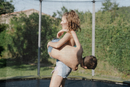 Horizontal photo of a blonde boy and a brunette girl fighting and playing on a trampoline on a green playground. The teenager and the child wear short jeans and they look happy, sm