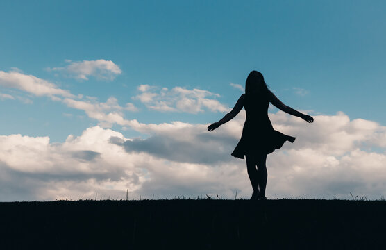 Silhouette of woman in a dress dancing against a blue sky with clouds.