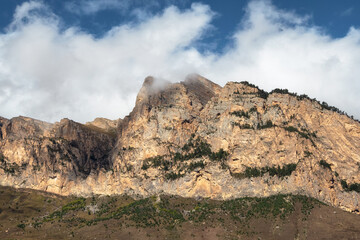 Beautiful long pointed rock is illuminated by the sun. Sunny mountain landscape with high sharp rockies under white clouds. Panoramic view of sharp rocks and peace top in blue cloudy sky.
