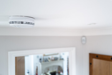 Shallow focus of a hard wired smoke detector seen in the downstairs hall, fixed to the ceiling. An...