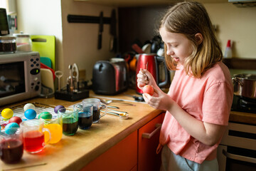 Pretty preteen girl using candle wax to dye Easter eggs at home. Child painting colorful eggs for Easter hunt. Kid getting ready for Easter celebration.