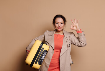 Lovely ethnic woman in stylish orange dress and beige coat, carrying a yellow suitcase, smiling at camera and gesturing with Ok hand sign, expressing agreement over beige isolated background