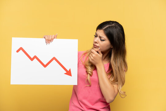 Young Russian girl isolated on yellow background holding a sign with a decreasing statistics arrow symbol and thinking