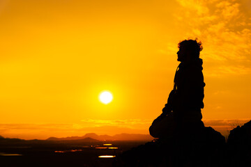 Silhouette of a man kneeling on a rock in prayer position at sunset time with the orange sky and the sun right in front of him. Concept of prayer. Copy space on the right