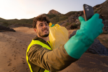 Volunteer smiles at the camera while taking a selfie with his mobile device. He carries a bag full of plastics collected from the beach. He is wearing gloves and a vest. He is on a beach at sunset.