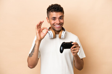 Fototapeta na wymiar Young Brazilian man playing with a video game controller isolated on beige background showing ok sign with fingers