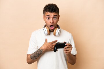 Fototapeta na wymiar Young Brazilian man playing with a video game controller isolated on beige background surprised and shocked while looking right