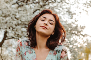 Portrait of charming pretty woman dressed flowery dress posing near apple cherry tree blossoms blooming flowers in the garden park in early spring nature. Fashion, girl model with black hair

