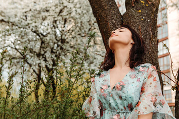 Portrait of charming pretty woman dressed flowery dress posing near apple cherry tree blossoms blooming flowers in the garden park in early spring nature. Fashion, girl model with black hair
