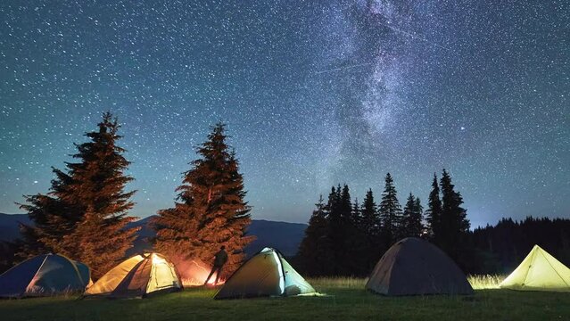 Night camping with five tourists illuminated tents. Hiker having a rest near campfire and forest, cooking dinner on fire. Timelapse of the starry sky with the Milky Way. Concept of tourism and nature.