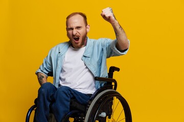 man in a wheelchair screams angrily, shows fist looks at the camera is not pleased, with tattoos on his hands sits on yellow background of the studio, the concept of health is person with disabilities
