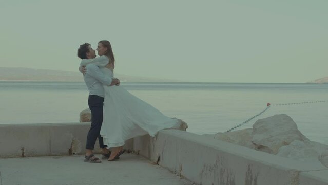 The bride and groom on the stone pier hugging and kissing against the sea at dawn. Lovely couple in wedding vacation.