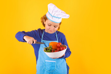 Kid cook hold plate with vegetables. Child chef cook prepares food on isolated studio background. Kids cooking. Teen boy with apron and chef hat preparing a healthy meal.