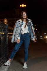 Attractive brunette woman in denim clothes and glasses on evening city background. Stylish girl on night city street