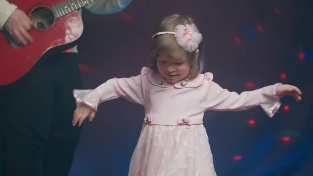 Little girl on stage in pink vintage dress, she dances, her father plays acoustic guitar. Color music is shining and smoke is billowing. Father Day. Performance on stage. Hobbies and creativity
