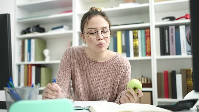 Young beautiful hispanic woman student writing on notebook eating apple at library university