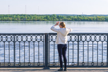 A young girl stands on the embankment and looks towards the river. A girl's walk by the river on a summer day. A blonde girl in a white windbreaker with her arms thrown back looks into the distance.