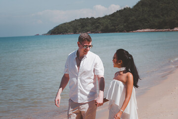 Couple walking on the beach romantic honeymoon vacation summer holidays romance .Young happy interracial couple walking on tropical beach smile holding around each other. Asian woman, Caucasian man.