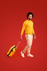 Serene curly 30s African-American guy in casual wear walking with yellow suitcase ready for adventure and holiday, isolated on red background, travel and vacation concept. Full length