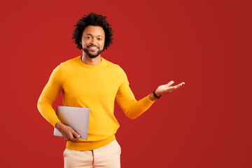 Cheerful handsome curly african-american guy hold laptop and point with palm aside at empty space...