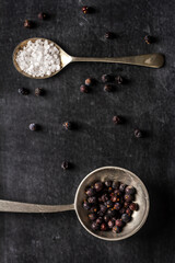 Two old spoons with juniper berries and salt on black background