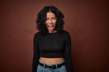 Young colombian curly hair woman isolated on brown background shouting very angry, rage concept, frustrated.