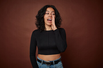 Young colombian curly hair woman isolated on brown background is saying a secret hot braking news and looking aside