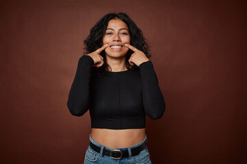 Young colombian curly hair woman isolated on brown background smiles, pointing fingers at mouth.