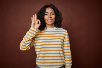 Young colombian curly hair woman isolated on brown background cheerful and confident showing ok gesture.