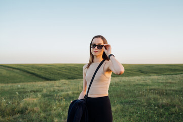 Portrait of a beautiful woman in casual clothes standing in a clean grassy field at sunset and posing for the camera.