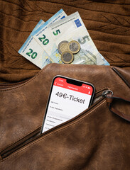 Aus einem geöffneten Reißverschluss einer braunen Handtasche schaut ein Smartphone heraus, auf dem gerade eine Fahrkarten-App mit einem 49€-Ticket darauf geöffnet ist. Im Hintergrund braun Strick - obrazy, fototapety, plakaty