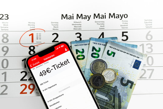 Auf einem Kalenderblatt vom Mai 2023 dessen 1. Mai mit einem Stift rot eingekringelt wurde, liegt ein Smartphone auf dem gerade eine Fahrkarten-App mit einem 49€-Ticket geöffnet ist und 49€ Bargeld, 