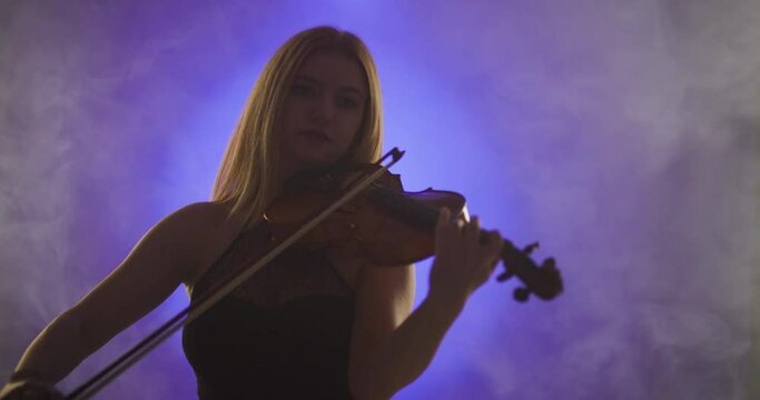 Beautiful blond woman playing violin against blue and violet spotlight with smoke background.