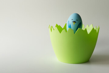 Niebieski wielkanocny kurczak zrobiony z jajka siedzący w zielonym naczyniu w kształcie skorupki jajka na jasnym tle. - obrazy, fototapety, plakaty