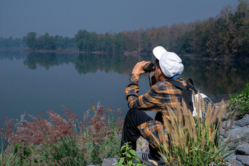 Asian boy in plaid shirt wears cap and has a backpack, holding a binoculars, sitting on ridge...