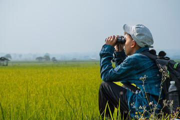 Fototapeta premium Asian boy in plaid shirt wears cap and has a backpack, holding a binoculars, sitting on ridge rice field of asian farmers to observe fish and birds on tree branches and on sky, soft focus.