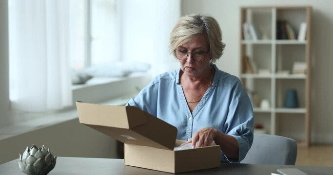 Attractive older woman, electronic commerce satisfied client opening cardboard box with ordered goods smile enjoy moment of purchasing items unpacking in parcel. Reliable transport company, e-commerce